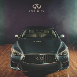 Infiniti Q50, sleek and shiny in a spotlight. Dark purple backdrop with large Infiniti logo. Professional automotive photography, showing the car's modern design and dark gray color.