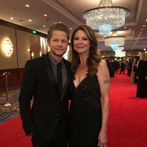 Matt Czuchry and Jane Leeves attending a glitzy Hollywood gala. The room is filled with sparkling chandeliers and red carpet. Matt's arm sports a prominent tattoo, Jane with a radiant smile.