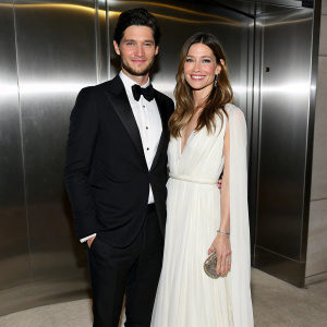 Ben Barnes in a black tuxedo, Jessica Biel in a flowing white dress. They stand at the MET Gala against a sleek silver backdrop.