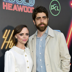 Christina Ricci and Adam Goldberg at a Hollywood event. Christina in a white blouse, Adam in a light jacket and striped shirt. They stand before a backdrop with neon logos.