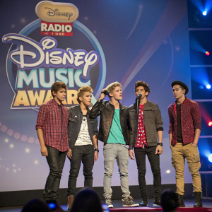 IM5 members on stage during the Radio Disney Music Awards. Dressed in trendy outfits. Radio Disney logo backdrop.