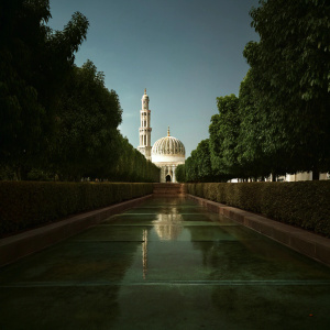 Sultan Qaboos Grand Mosque, located in Muscat, Oman, features a vast white architectural design with a prominent dome and towering minaret. The mosque is set within a lush plaza, adorned with reflective pools and meticulously manicured hedges.