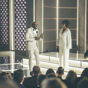 DJ Black Coffee and Esona Maphumulo performing at the Grammy Awards Premiere Ceremony. The stage is brightly lit with purple patterns. Esona Maphumulo in a white suit accepts an award as the audience watches.