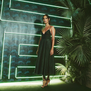Adria Arjona at the star-studded Comic-Con event. Wearing a black halter dress with high heels. Neon pink and yellow backdrop with Comic-Con branding.
