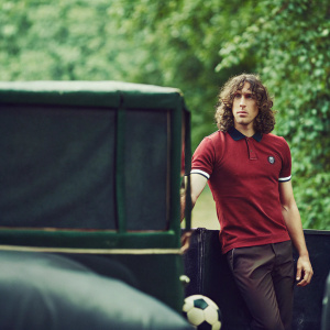 Carles Puyol, wearing a red polo shirt with the number 18, standing solo against a sleek black backdrop. Long curly hair framing his focused expression.