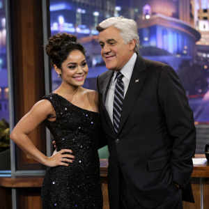 Vanessa Hudgens, with elegant updo and sparkling black dress, next to Jay Leno in a talk show studio. Formal attire, warm lighting, urban backdrop.