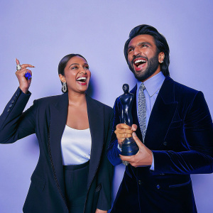 Sonakshi Sinha and Ranveer Singh, joyfully posing at the Filmfare Awards. Sonakshi in a black blazer over a white top. Ranveer in a black tuxedo with a patterned tie. White backdrop with festival logos.
