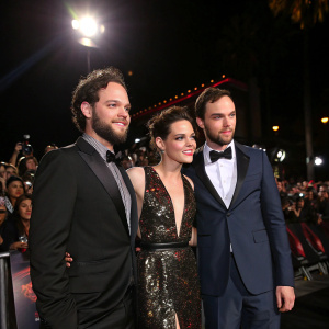 Drake Doremus, Kristen Stewart, and Nicholas Hoult at a Hollywood premiere. Dark stage with spotlight on them, surrounded by fans and paparazzi.