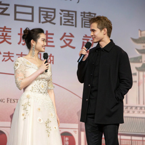 Hayden Christensen and Liu Yifei attending the Shanghai Film Festival premiere. Liu Yifei in elegant white gown with intricate embroidery and floral details, addressing the crowd with microphone. Christensen in casual attire with black jacket. Chinese characters and traditional architecture backdrop.