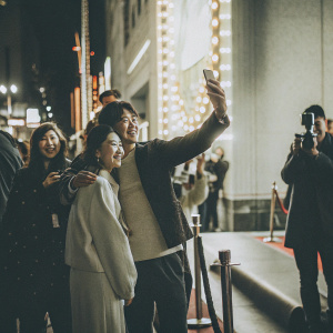 Lee Sang-Yun and Noh Young-Hak at a Hollywood event, in casual outfits, posing for selfies with fans. Neon lights, bustling crowd