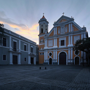 Golden hour photography of St. Agustin Church in Intramuros, Manila. The church is a warm-toned structure with intricate orange and white detailing. Multiple arched windows and a tall bell tower dominate the scene.