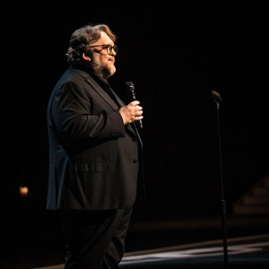 Guillermo del Toro, in a black suit, giving an acceptance speech. Spotlight illuminates him on a dimly lit stage.