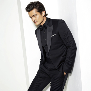 Orlando Bloom in a sleek black tuxedo, posing for a close-up shot. Stark white background