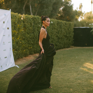 Bella Hadid in elegant black dress, posing seriously at gala. White backdrop with sponsors' logos