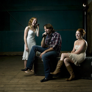 Snapshot from 'The Voice'. Miley Cyrus in a white dress, Aaron Gibson in plaid shirt, Ali Caldwell with red hair and pale dress. Candid moment on stage