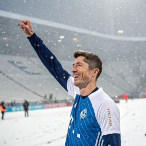 Robert Lewandowski, a football star, grinning and raising his arm in celebration. Wearing a blue and white jersey, amidst a snowy stadium scene.
