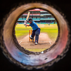 Cricket match photo of Eoin Morgan. England captain, batting stance. Blue jersey with white logo, dark protective gear. Cricket field with grassy outfield and worn-out batting area.