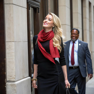 Amber Heard, blonde hair, black dress, red scarf, standing outside with a building backdrop. Man with big smile standing beside her.