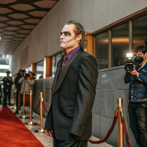 Picture of Joaquin Phoenix as Joker at film premiere. Dressed in black suit with purple shirt, serious expression. Background is bustling red carpet with flashing cameras.