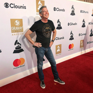 Elvis Duran, a friendly host, in a casual outfit: black graphic t-shirt and jeans, stands confidently at the Grammy Awards red carpet. Logos of various sponsors displayed on white backdrop.