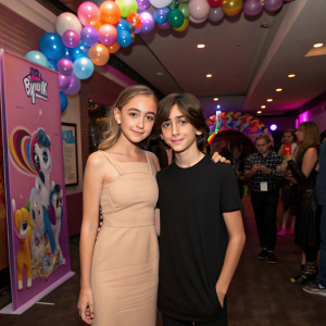 Gabrielle Elyse and Aidan Gallagher attending a My Little Pony themed party. Gabrielle in a stylish beige dress, Aidan in a black t-shirt. Crowded venue with My Little Pony decorations.