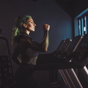 Green-haired She-Hulk flexing her muscles in a sleek combat suit. Training in an advanced tech gym with futuristic equipment and a neon-lit blue backdrop.