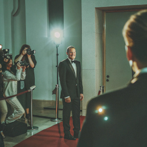 Woody Harrelson at a glitzy gala. Wearing a black tuxedo. Red carpet background, paparazzi in the corner