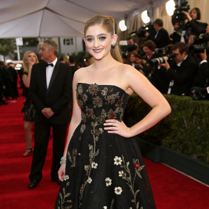 Willow Shields at a high-profile awards show. Elegant black dress adorned with intricate floral details. Wears a red carpet gown. Glowing smile. Formal setting with celebrities and flashing cameras.