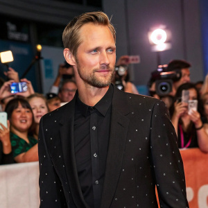 Alexander Skarsgård at a film festival. Wearing a black suit, stubble, and short hairstyle. Crowded venue with flashing cameras.
