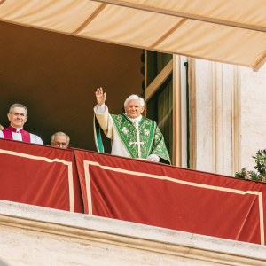 Pope Benedict XVI waving to the crowd. He is in a green papal robe adorned with crosses and other religious symbols. Surrounded by other clerics in white robes. A bustling outdoor event with many onlookers.