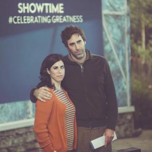 Sarah Silverman and Sacha Baron Cohen at the Showtime Awards. Sarah, in an orange sweater and striped shirt, beams at the camera. Sacha, in a dark zippered jacket, has a serious expression. They stand against a vibrant blue backdrop with the Showtime logo and the hashtag #CelebratingGreatness