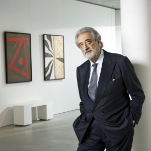 Placido Domingo, the renowned opera singer, poses in a minimalist studio setting. He wears a dark suit with a subtle patterned tie. Serious and composed demeanor. Modern art pieces adorn the background.