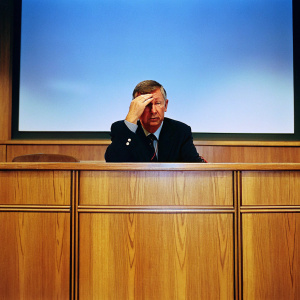 Sir Alex Ferguson, intense look. Dark blazer. Gray hair. Hand on forehead. Conference room background