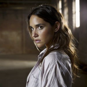 Adria Arjona in a black and white photo, dark brunette waves framing her serious face. Wearing a light-colored, button-up shirt with visible texture. The background is blurred for emphasis on her attire and expression.