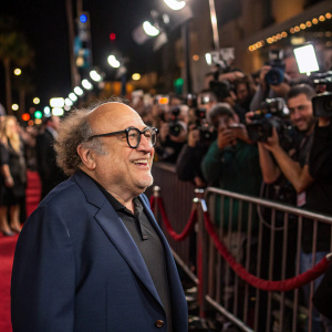 Danny DeVito, with his signature curly hair and glasses, at a bustling Hollywood event. He's dressed in a navy blue suit. Surrounded by flashing cameras and fans.