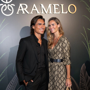 Julio José Iglesias Jr. and Charisse Verhaert attending the Aramelo brand launch party. Formal attire, black suit and patterned dress, posing in front of large Aramelo logo backdrop.