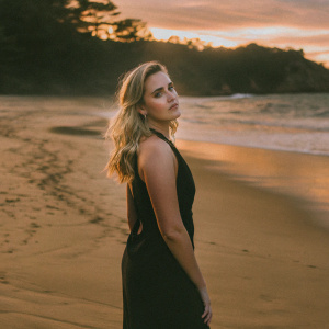 Maggie Grace, blonde hair, elegant makeup, standing alone on a sandy beach at sunset. Wearing a black dress.