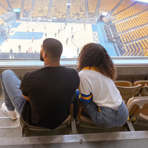 Drake and Skylar Diggins at an NBA game. Drake in a casual black t-shirt and jeans, Skylar in a sports jersey, sitting courtside