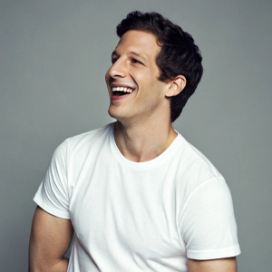 Andy Samberg, with a big smile, wearing a white t-shirt. Close-up shot, simple grey background