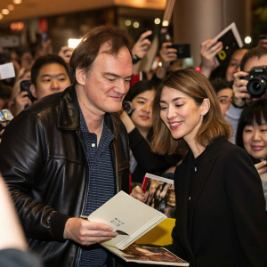 Quentin Tarantino and Sofia Coppola at a bustling book signing event. Both are dressed casually. A crowd of eager fans surround them.