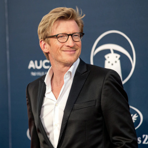 David Wenham, a friendly-faced caucasian male with short blonde hair and glasses, stands in front of a dark blue backdrop adorned with a logo. He wears a sleek black suit jacket over a crisp white shirt.