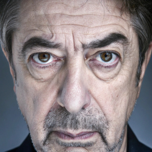 Joe Mantegna, the actor, photographed up close. His grizzled face, with a mix of gray and brown hair, exudes seriousness. Dark background emphasizing facial features.