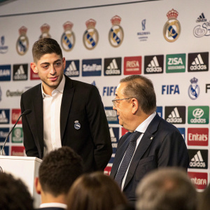 Real Madrid's Federico Valverde and a distinguished gentleman in a black suit and glasses stand before a bustling crowd at a high-profile press conference. The backdrop showcases numerous logos, such as FIFA.com and WANDA, amid a sophisticated indoor setting.