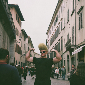 Johnny Bravo lookalike, flexing his muscles in a crowded street. His outfit includes a black t-shirt, and he's sporting a big, blonde, pompadour hairstyle.