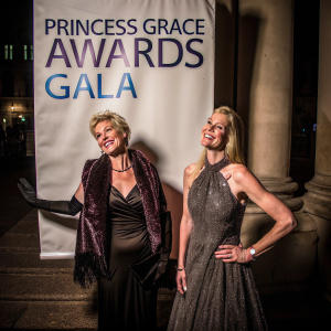 Charlene of Monaco and Giselle Paulson at the star-studded Princess Grace Awards Gala. They are posing in front of a large white backdrop adorned with blue logos and text.