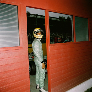Lewis Hamilton in a Formula 1 garage, dressed in his Petronas McLaren Racing suit and helmet. Crowds cheering outside.