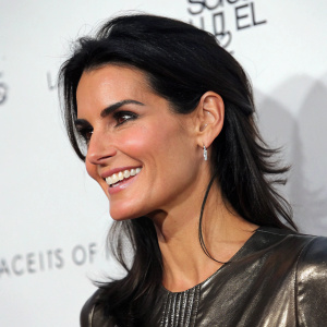 Angie Harmon, a smiling yet slightly melancholic actress, posing in a close-up shot. Wearing a sleek, shiny top. Dark, layered hair framing her face. The backdrop is a plain white wall with subtle text.