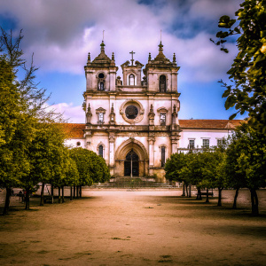 A breathtaking photograph of the ancient sanctuary in Alcobaça, Portugal. Grand façade with a central archway, surrounded by statues and intricate details. Constructed from luminous stone, it stands as a symbol of classic European elegance. The scene is framed by a vibrant blue sky dotted with fluffy white clouds. Situated in an open courtyard with flat sandy ground, the architecture unfolds in multiple tiers of ornamentation, centered around the majestic entrance.