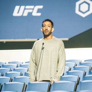 Dan Ige, UFC fighter, at a press conference. Gray t-shirt, standing in front of bright UFC logo backdrop. Formal event setting.