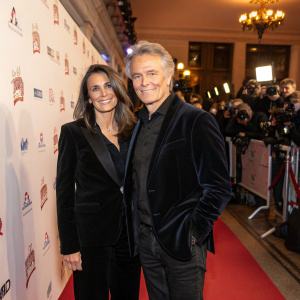 Danièle Dubosc and Franck Dubosc attend the opening night of 'Alibi.com 2'. The venue is The Grand Rex, bustling with photographers and fans. White backdrop with event sponsor logos. Both wear tailored dark velvet blazers.